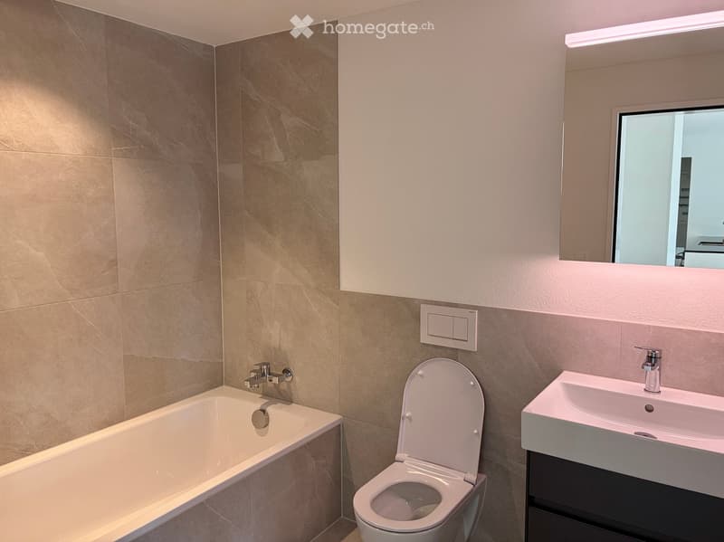 Modern bathroom with beige marble tiles, a bathtub, toilet, sink, and a large mirror above the sink.