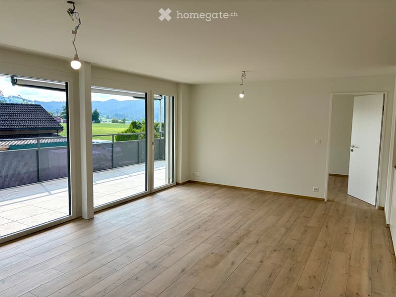 Empty room with wooden flooring, large sliding glass doors leading to a balcony with a scenic view of a green field and mountains.