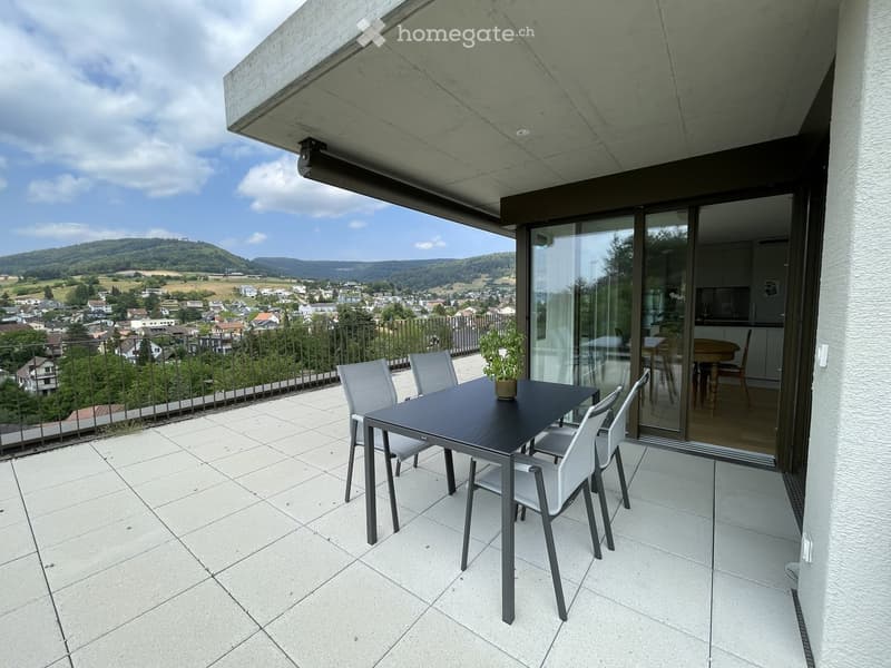 Balcony with a modern black table, four white chairs, and a small potted plant, overlooking a scenic town and countryside view.