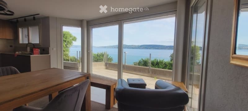 Bright, modern living space with large windows offering a scenic view of a lake. Features a wooden dining table with chairs, a leather sofa, and a small bench near the sliding glass doors leading to a balcony.