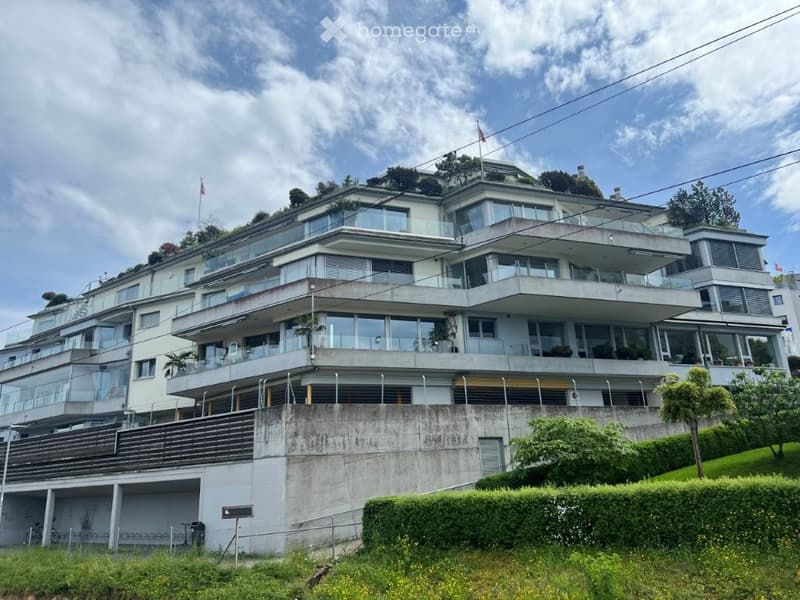 Modern multi-story building with large glass windows and balconies, situated on a raised ground floor with a landscaped garden in front.