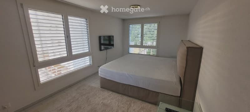 Modern bedroom with a large bed, headboard, and a flat-screen TV mounted on the wall. The room features large windows with blinds and a neutral color palette.
