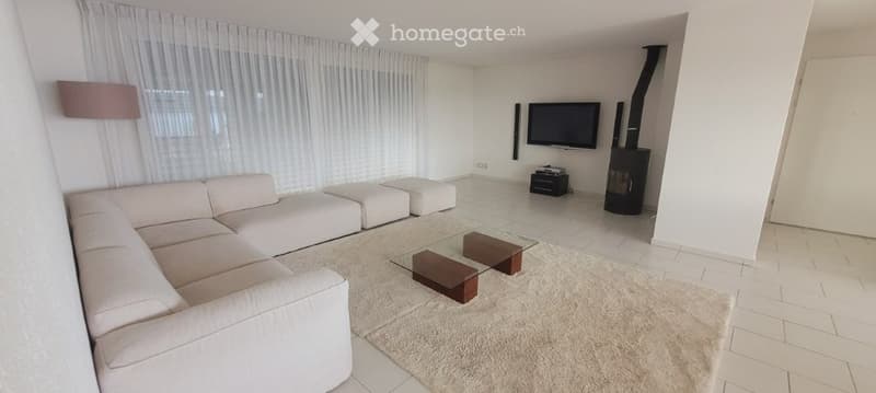 Modern living room with white walls, large windows with sheer curtains, a white sectional sofa, ottomans, a glass coffee table, a floor lamp, a television, and a fireplace.