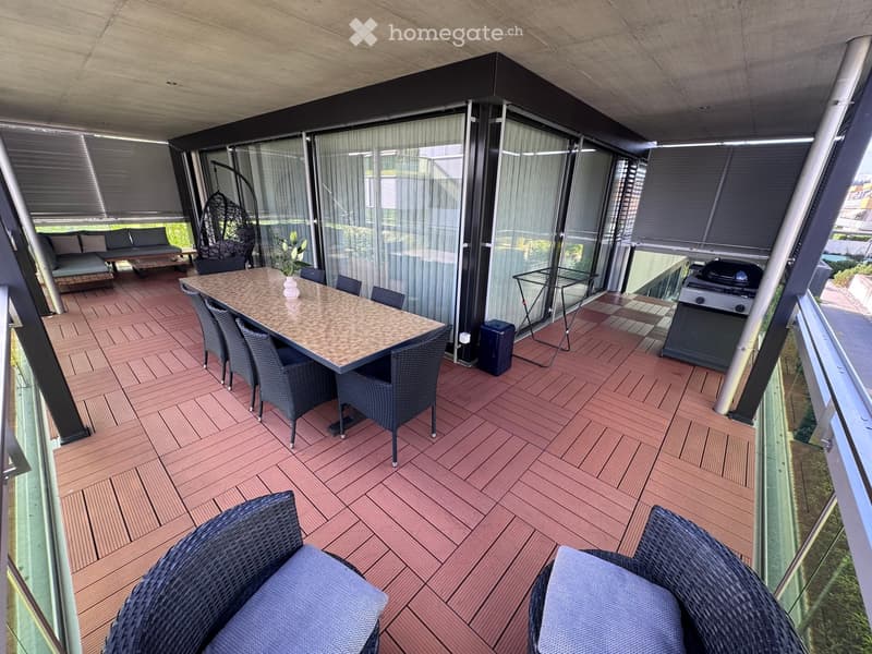 Balcony with wooden decking, glass walls, a dining table with chairs, a sofa, a hanging wicker chair, and a grill.
