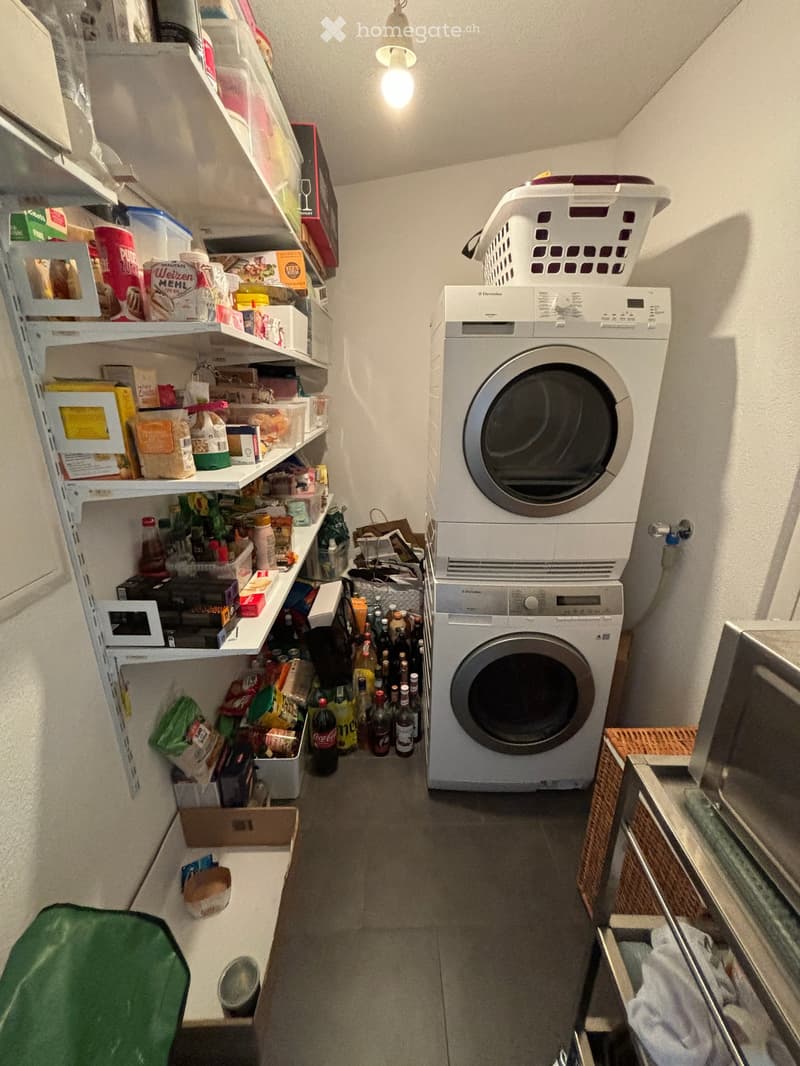 A small, well-lit room with a washing machine and dryer stacked vertically, shelves filled with food items and household supplies, and a laundry basket on top of the dryer.