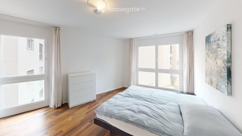 Bright bedroom with wooden flooring, large windows with curtains, a bed with grey bedding, a radiator, and a modern abstract artwork on the wall.