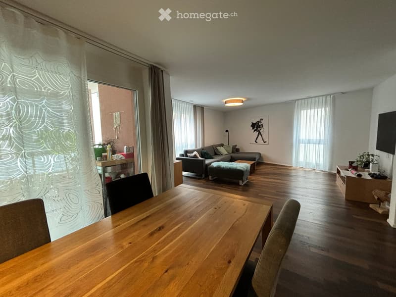 Open-plan living and dining area with wooden flooring, large windows, a grey sofa, coffee table, ottoman, dining table with chairs, and a TV screen.