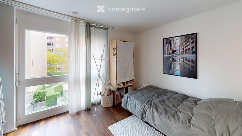 Modern bedroom with wooden flooring, large window with view, bed with grey duvet, bookshelf, picture frame, and coat rack.