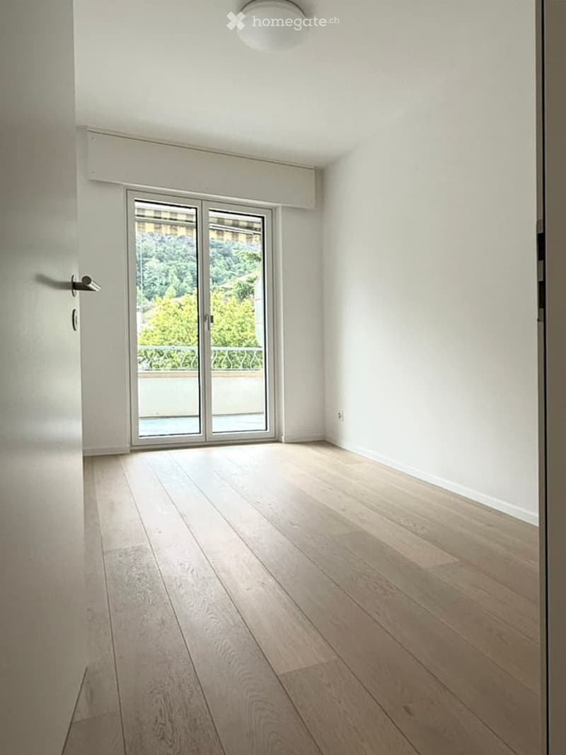 Empty room with light wooden flooring, white walls, and a large sliding glass door leading to a balcony with a view of greenery.