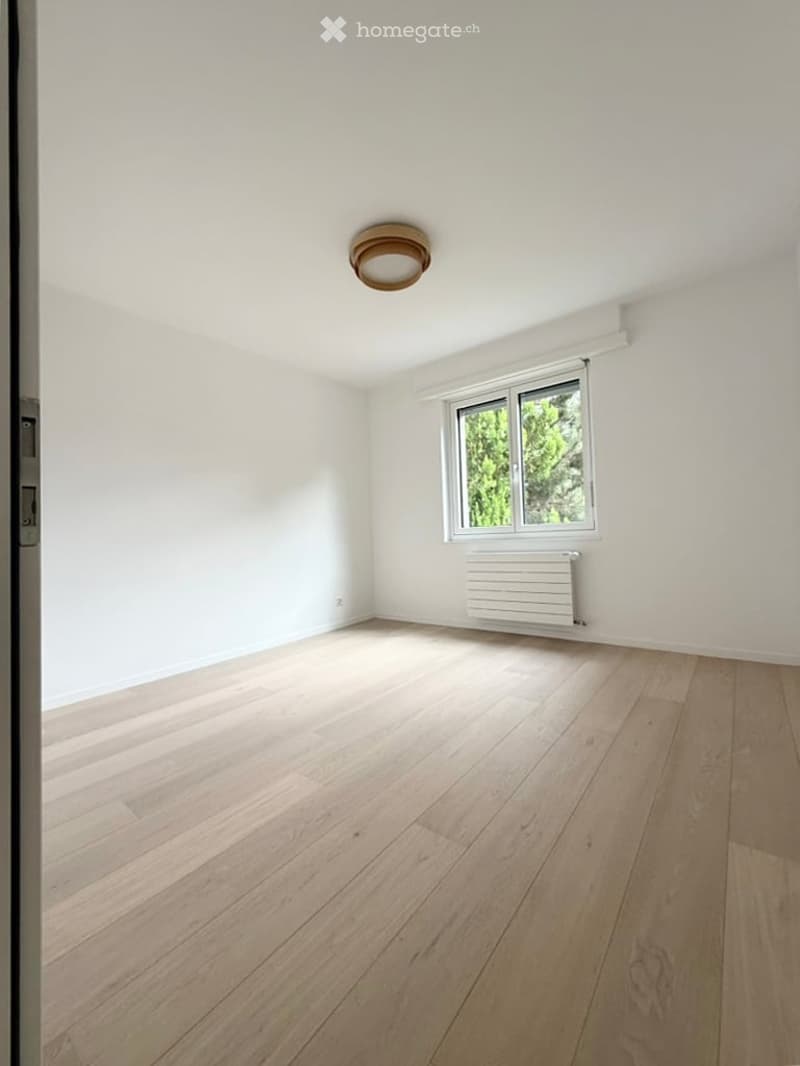 Empty room with light wooden flooring, white walls, a single window with white frame, and a white radiator below the window. The ceiling features a simple round light fixture.