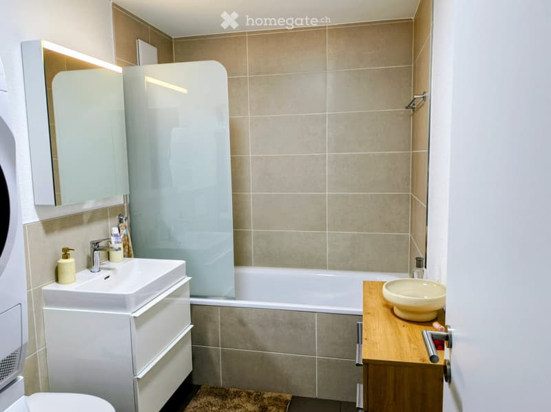 Modern bathroom with beige tiles, white sink, bathtub, and a wooden shelf holding a bowl. Features a large mirror with built-in lighting and a glass shower enclosure.