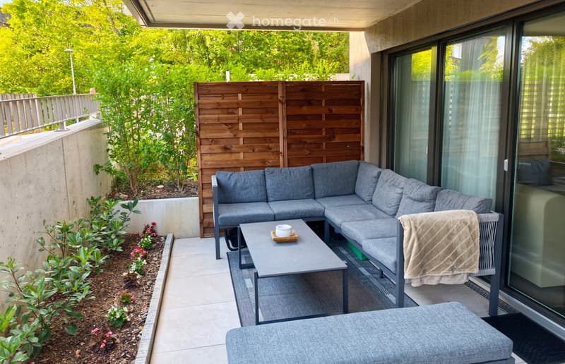 Modern balcony with grey sectional sofa, coffee table, and a small chair with a blanket draped over it. Wooden lattice screen and small garden area with plants and flowers.