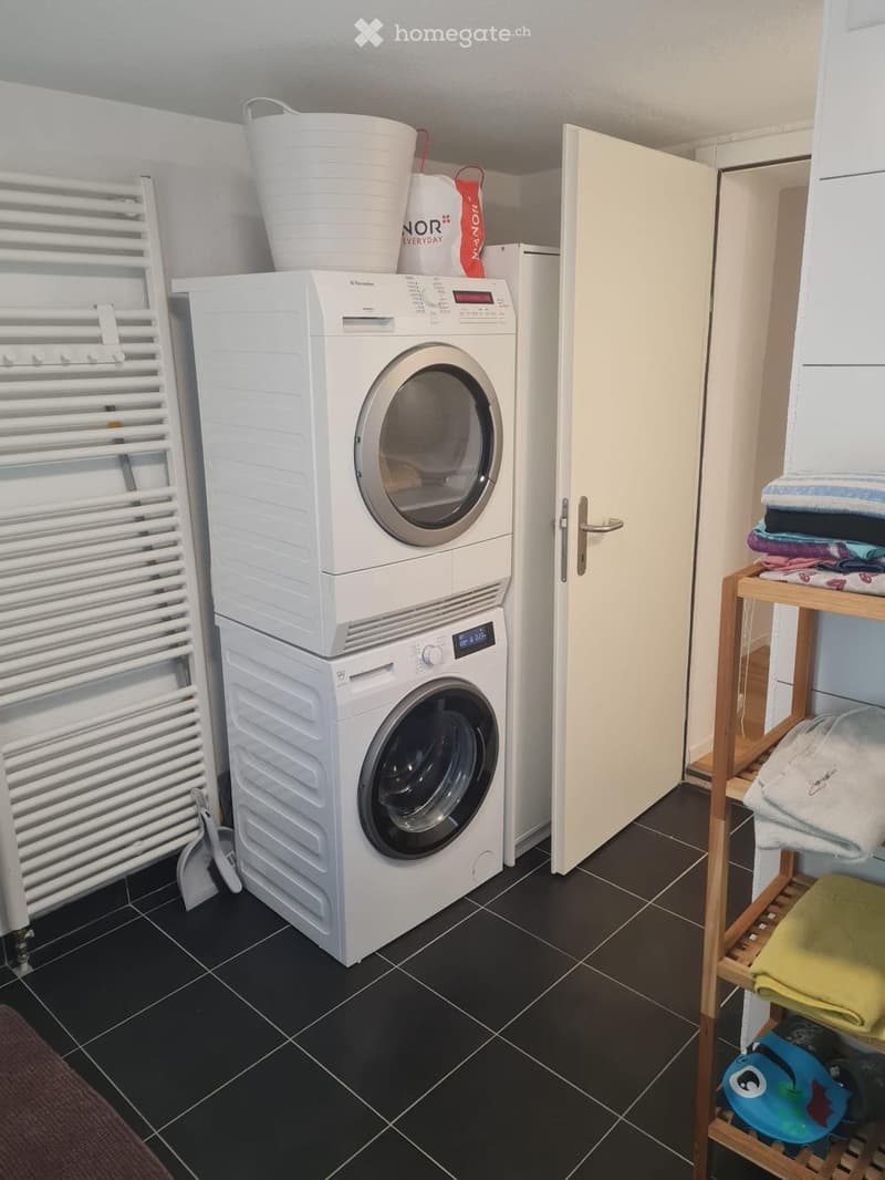 A compact bathroom with a stacked washing machine and dryer, a towel rack, and a shelf holding towels and other items.