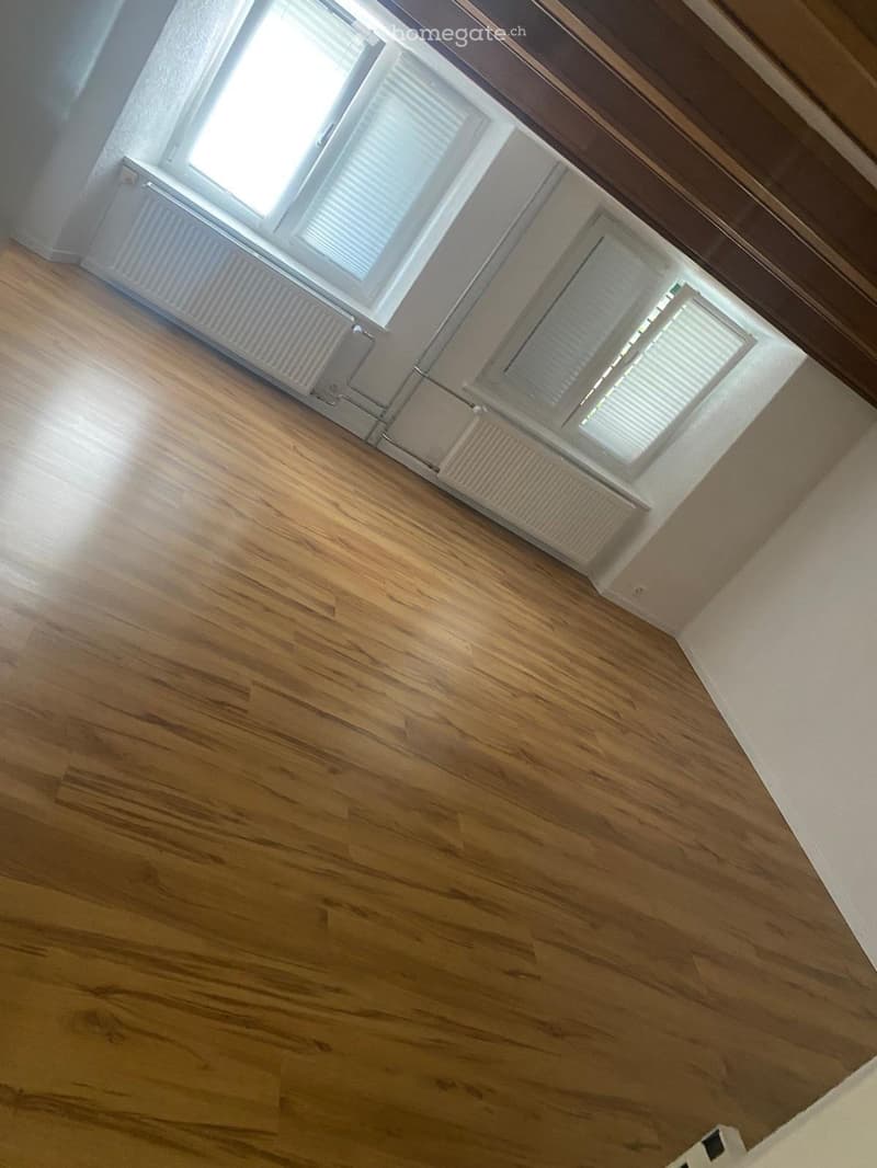 Empty room with wooden flooring, white walls, and large windows with blinds. Radiators are visible along the walls.
