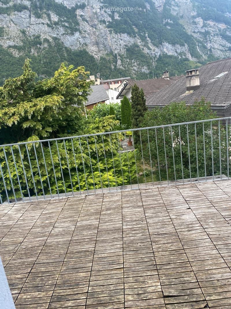 Wooden deck with metal railing, overlooking a lush green area with trees and distant mountains.