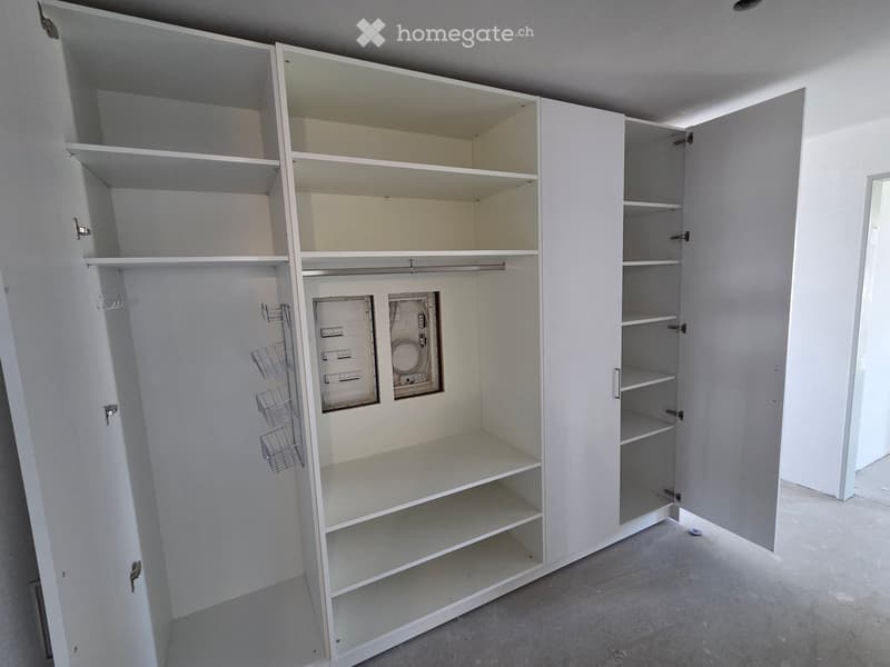 Empty room with white built-in wardrobes and shelves, featuring a visible electrical panel and storage baskets.