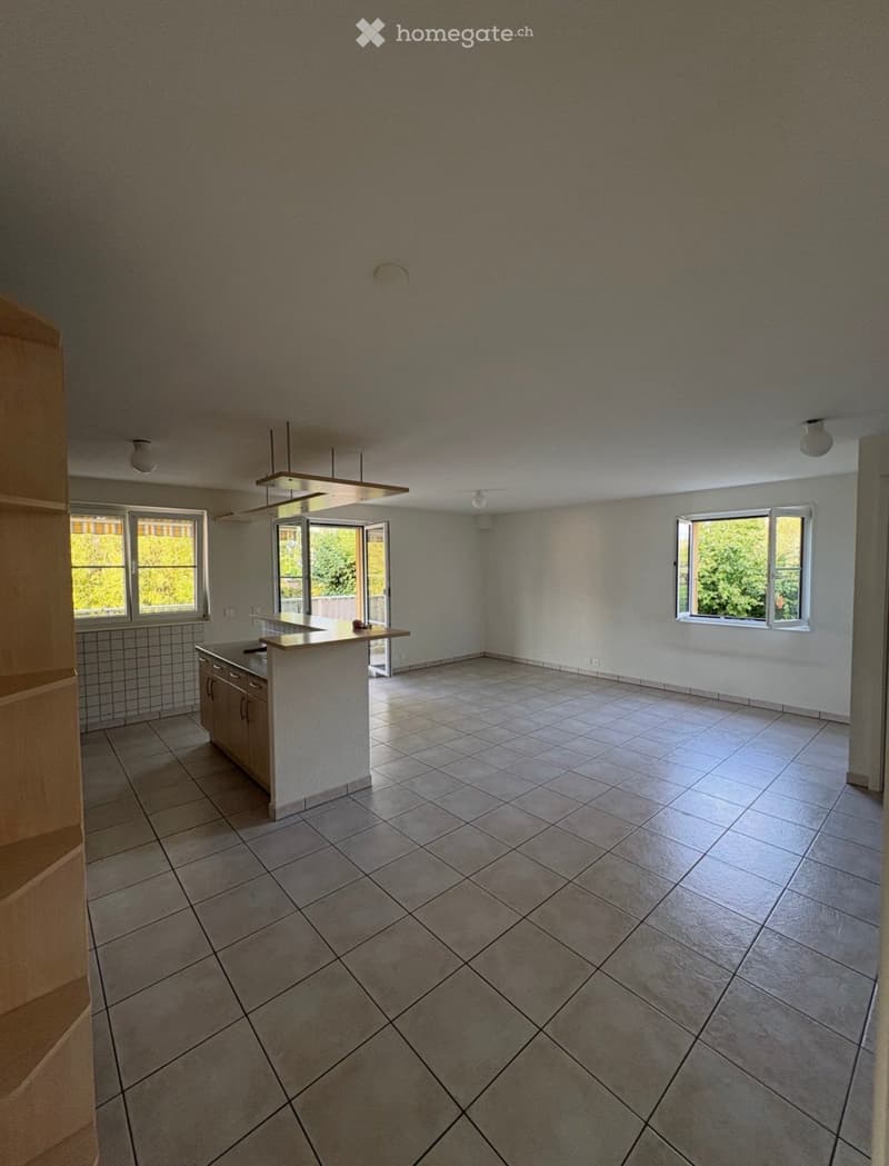 Empty kitchen with tiled floor, white walls, and a small countertop with cabinets. Three windows provide natural light, and a sliding door leads outside.