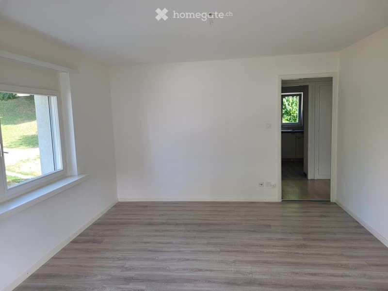 Empty room with light-colored walls, wooden flooring, and a large window with a view of the outdoors. A doorway leads to another room with visible furniture.