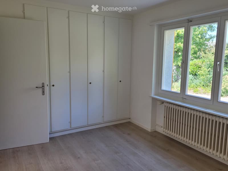 Empty room with light wooden flooring, white walls, built-in wardrobes, and large windows with a radiator below.