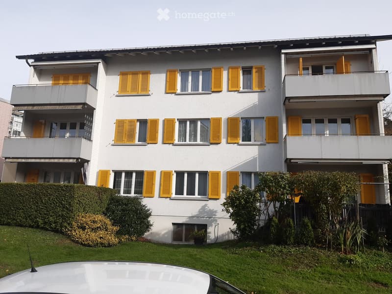 A three-story white building with yellow shutters, balconies, and a well-maintained garden in front.