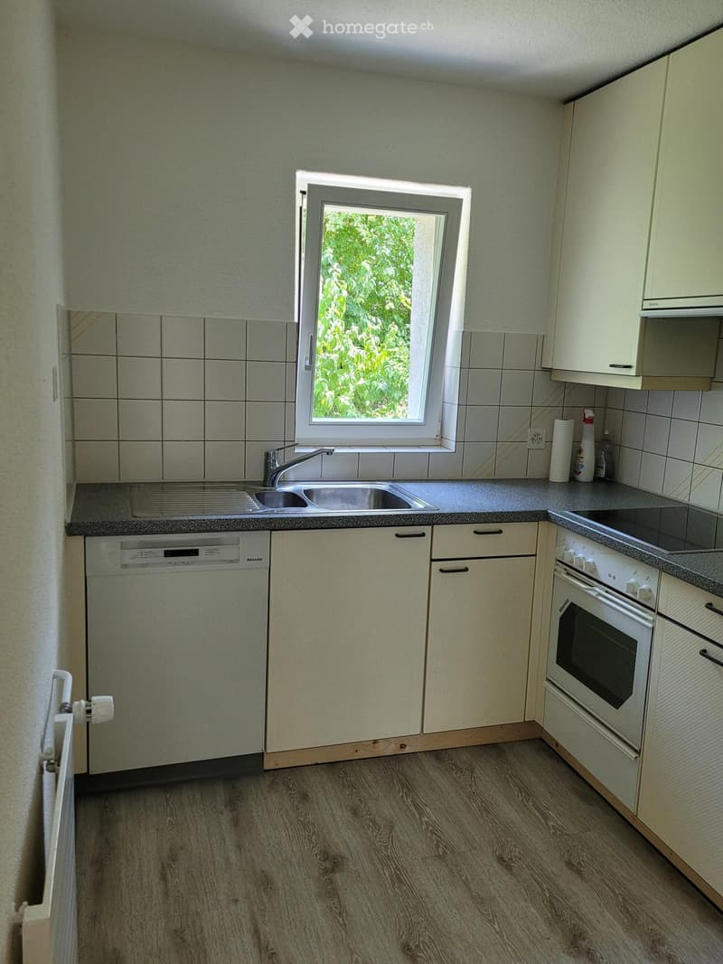 Modern kitchen with light-colored cabinets, a stainless steel sink, a dishwasher, and an oven. The countertop is tiled, and the floor is wooden.