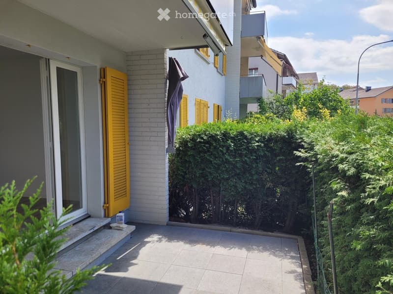 Balcony with tiled floor, white brick wall, yellow shutters, and a view of greenery and neighboring buildings.