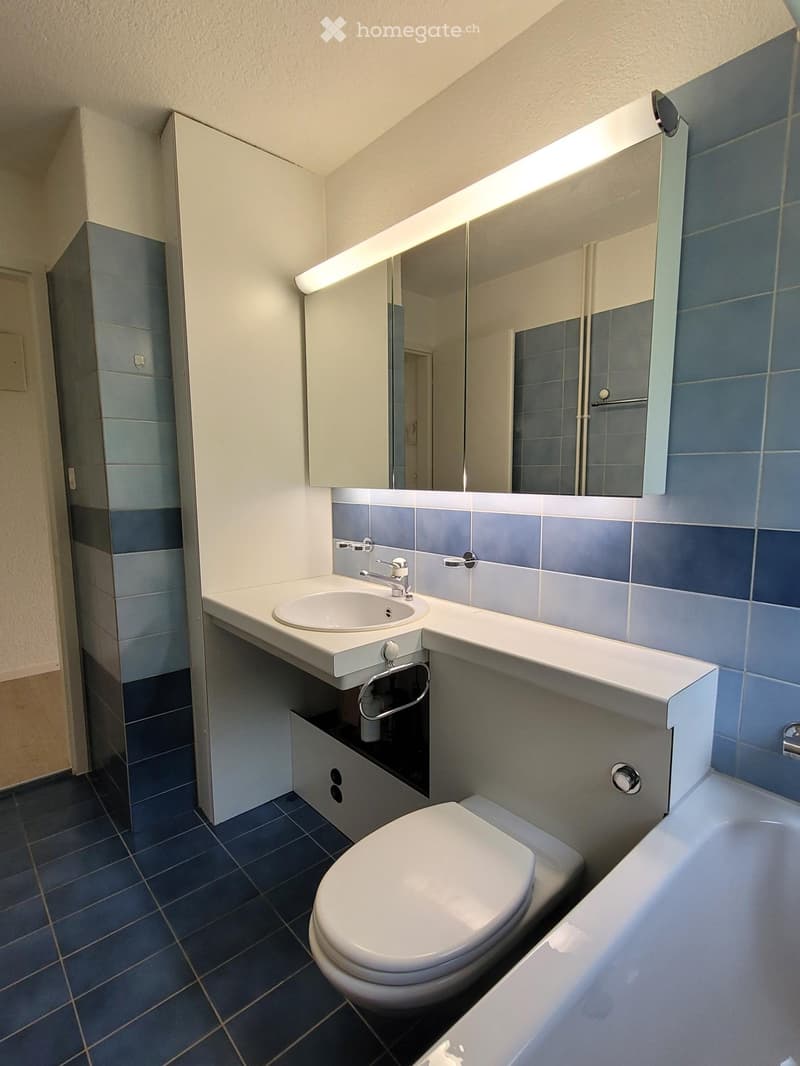 Modern bathroom with blue tiles, white sink, toilet, bathtub, and mirrored cabinet with built-in lighting.