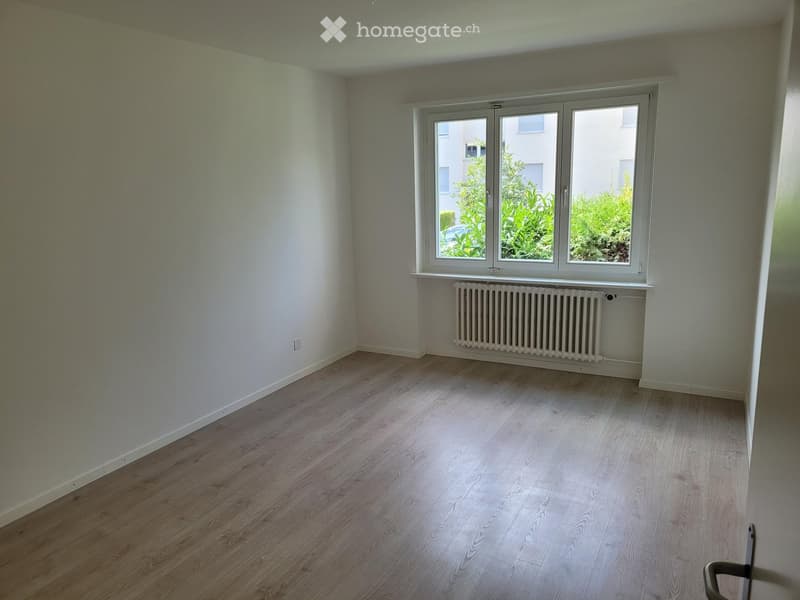 Empty room with light wooden flooring, white walls, and a large window with a radiator below it.