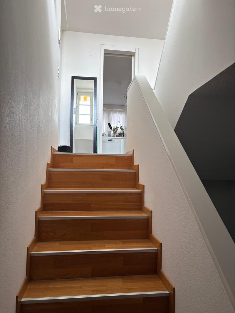 Wooden staircase with white railings, leading to a room with a window and a small kitchen area visible in the background.