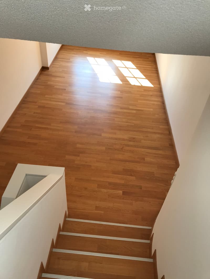 Wooden staircase leading up, with wooden flooring and white walls, natural light from a skylight above.