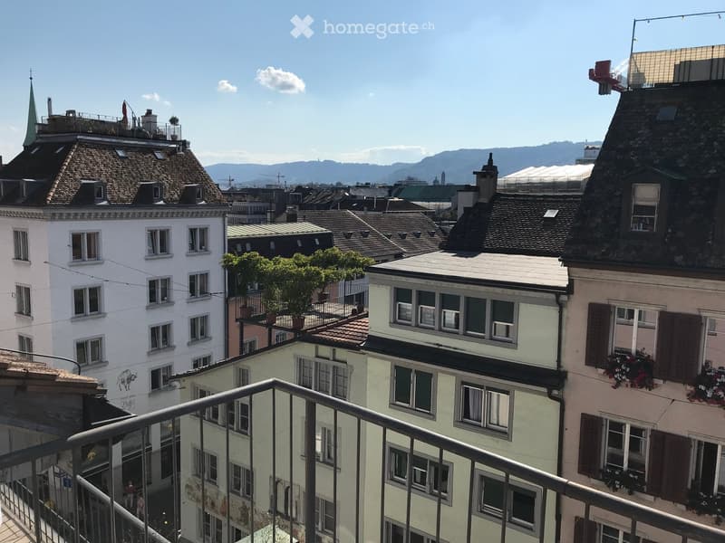 Balcony overlooking a cityscape with multiple buildings, including a white building with many windows, a green-roofed building, and a pink building with shutters and flower boxes. The background shows a clear blue sky, distant mountains, and a church spir