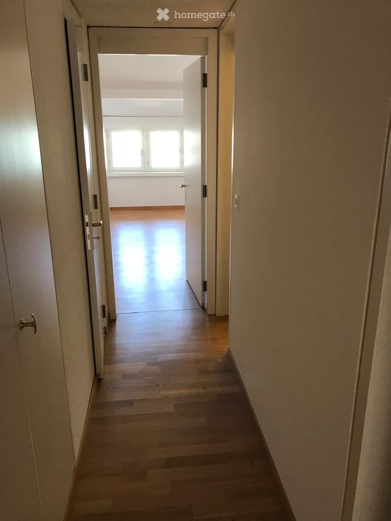 Empty hallway with wooden flooring, white walls, and a door leading to a room with a window.