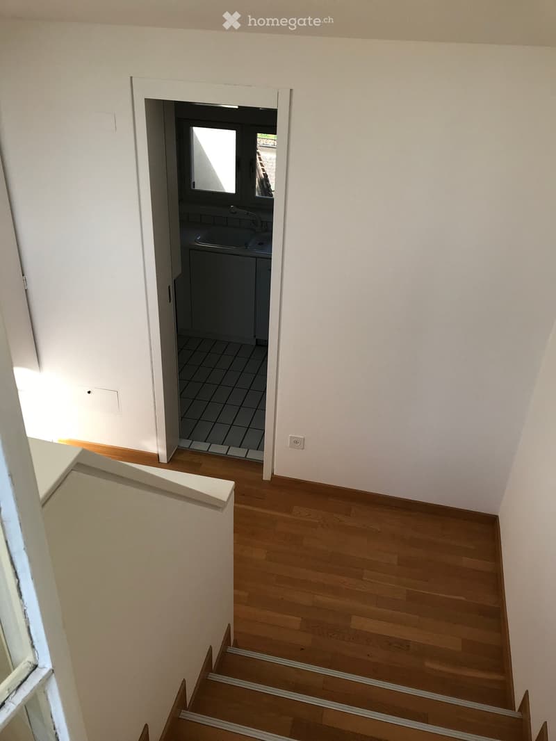 A narrow hallway with wooden flooring and white walls, leading to a small bathroom with a sink and tiled floor.