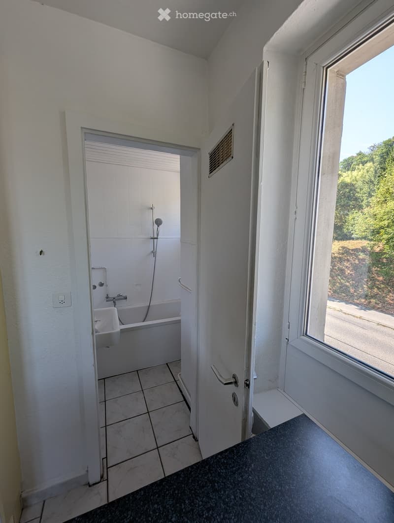 Bathroom with a bathtub, showerhead, and faucet. The room has tiled floors and walls, with a window offering a view of greenery outside.