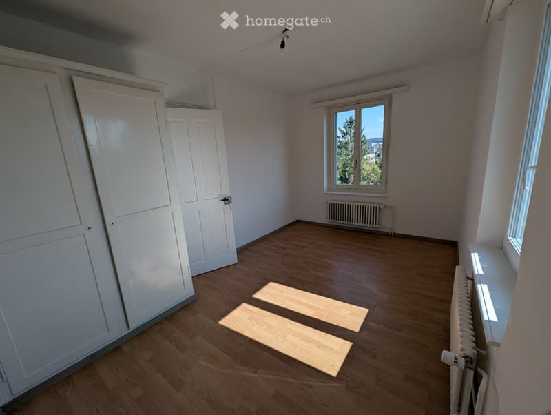 Empty room with wooden flooring, white walls, and large windows offering a view of trees and buildings. There are two white doors on the left side and radiators along the walls.
