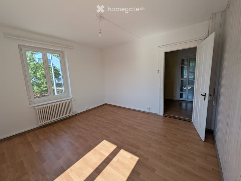 Empty room with wooden flooring, white walls, a window with a radiator below, and an open door leading to another space.