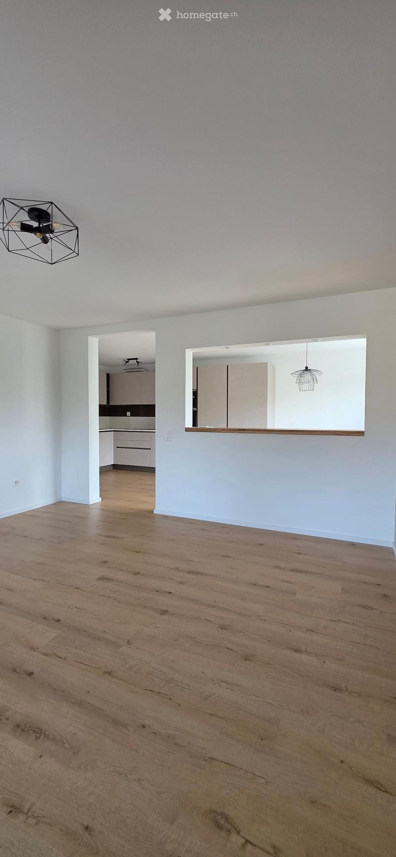 Empty room with light-colored walls and wooden flooring, featuring a modern geometric light fixture and an open doorway leading to a kitchen area.