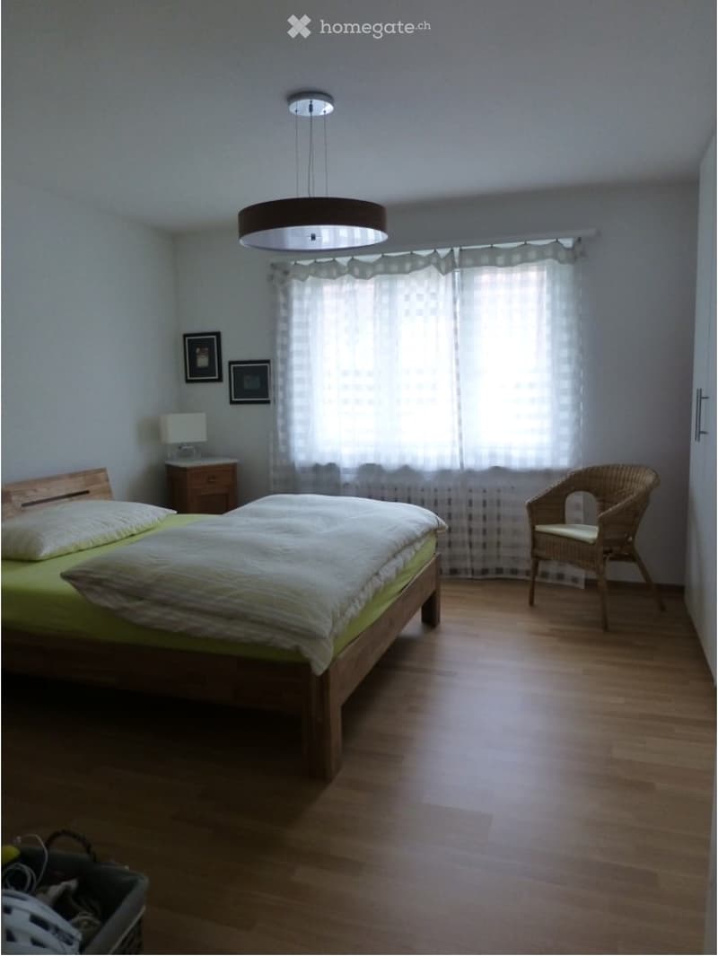 Bright bedroom with wooden flooring, large window with sheer curtains, bed with green bedding, nightstand with lamp, wicker chair, and two picture frames on the wall.