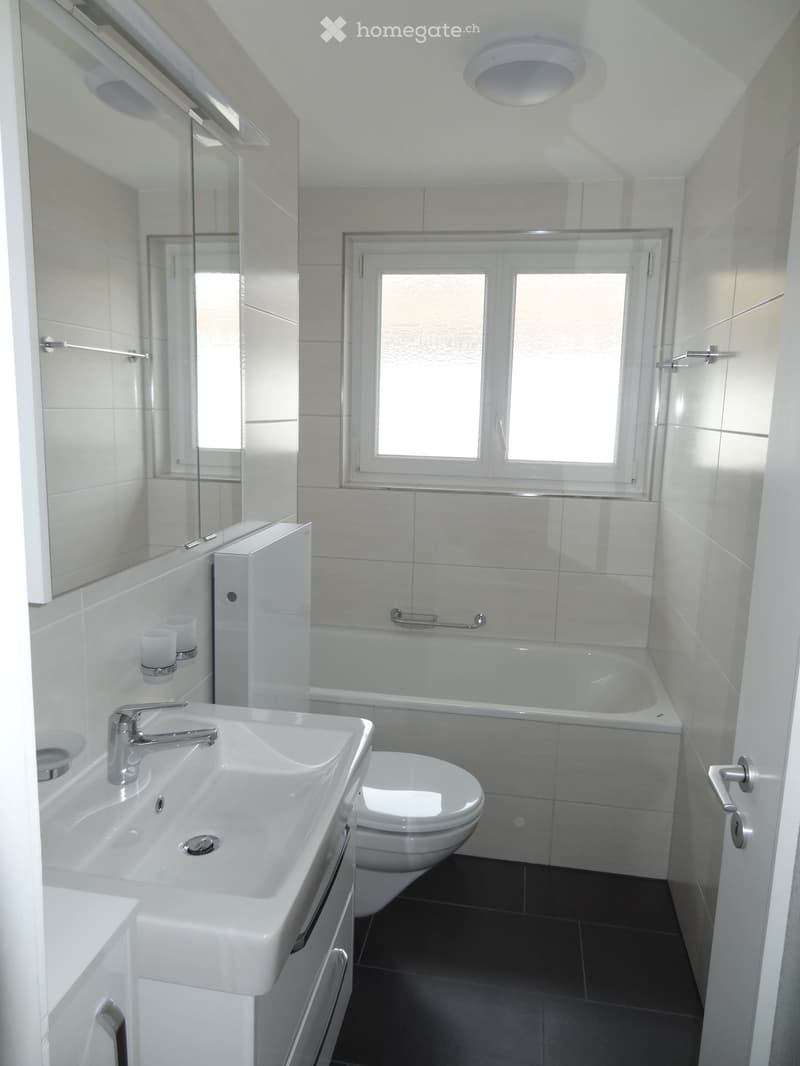 Modern bathroom with white tiles, a large sink, a toilet, a bathtub, a mirror, and a window letting in natural light.