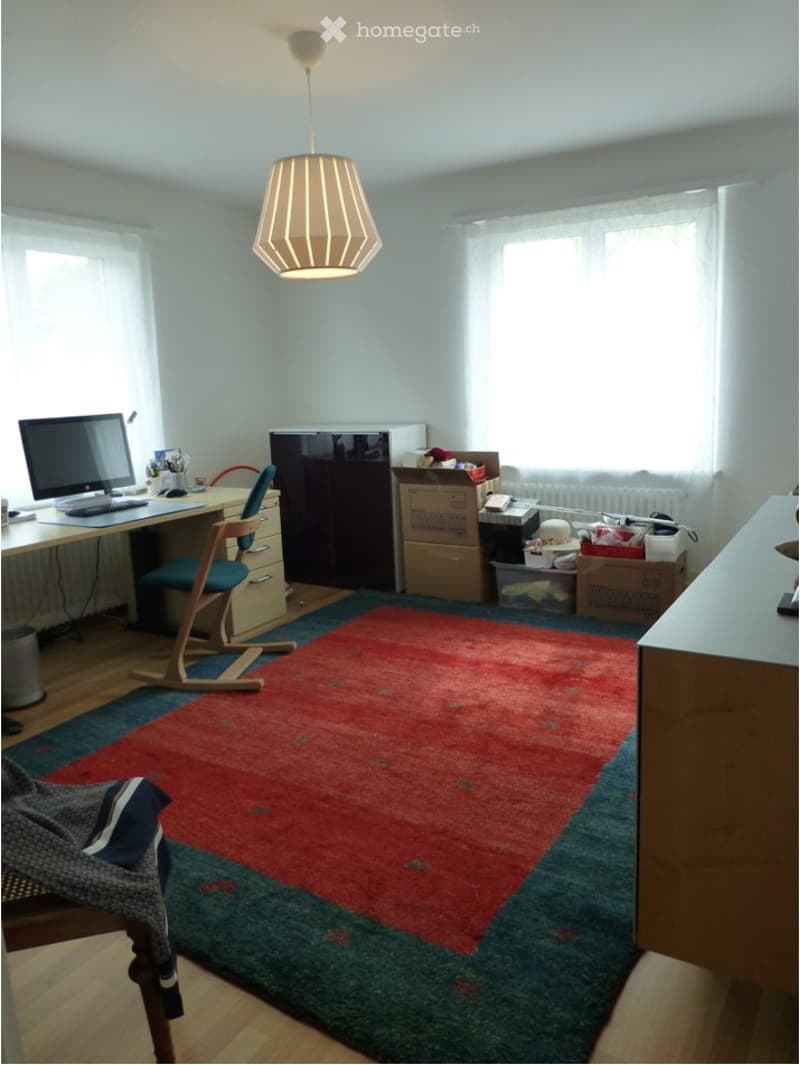 A furnished room with a desk, chair, computer, lamp, refrigerator, and a colorful rug on the floor.