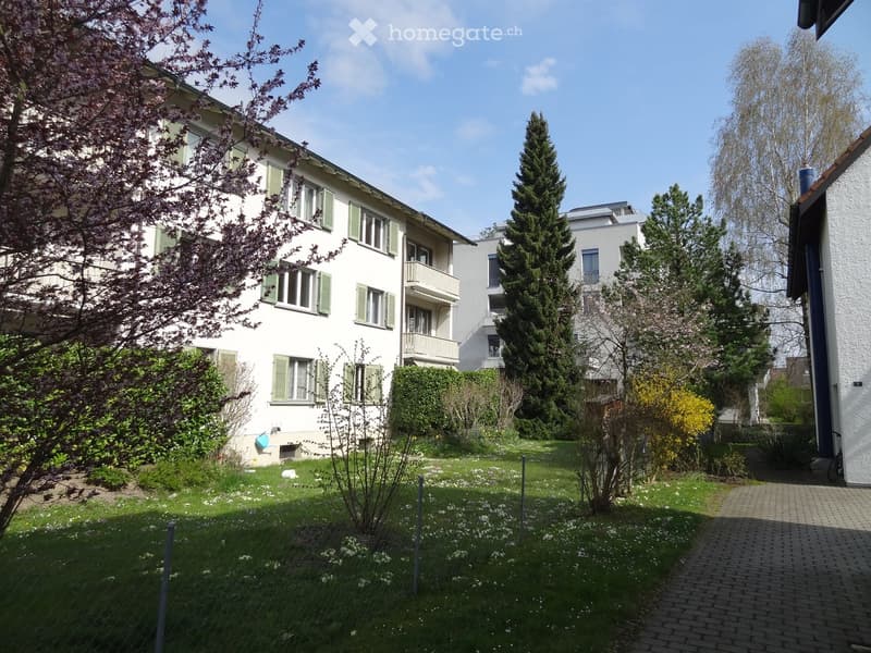 Well-maintained garden area with green grass, flowering plants, and trees, adjacent to a multi-story residential building with balconies and green shutters.