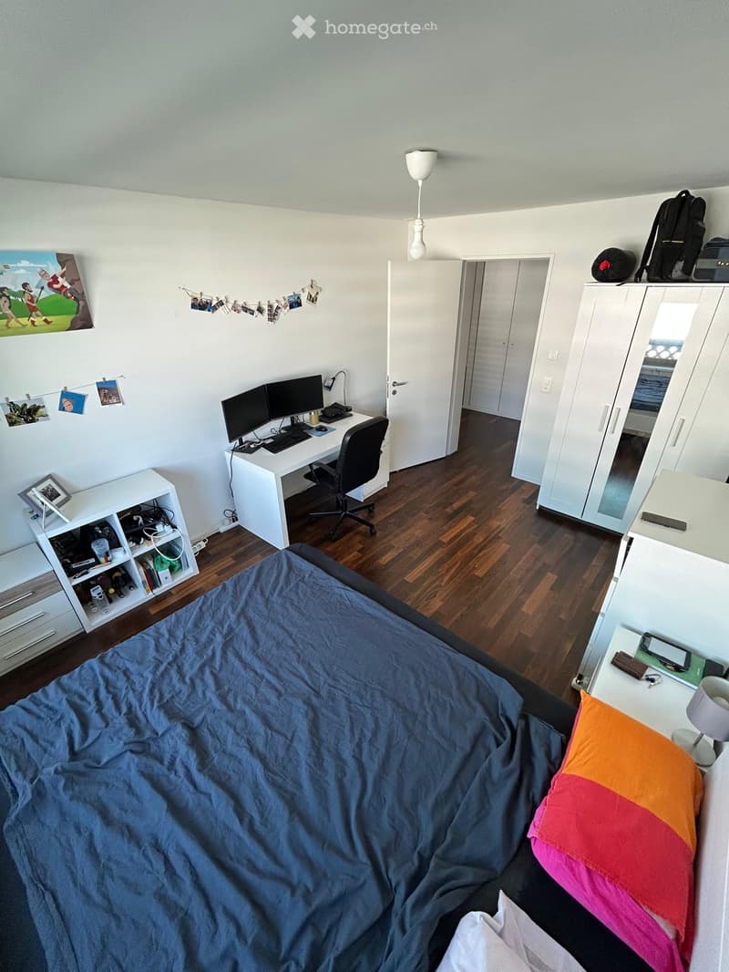 A well-lit bedroom with a bed, desk, and bookshelf. The room features a wooden floor, white walls, and a large window with a view.