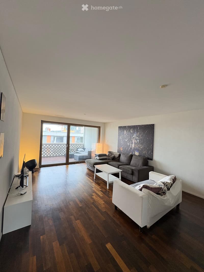 Modern living room with wooden flooring, large sliding glass doors leading to a balcony, a grey sectional sofa, a white coffee table, a white armchair, and a TV on a stand.