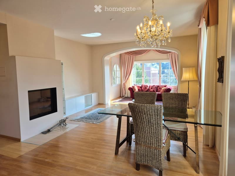 Elegant living and dining area with wooden flooring, a glass dining table with wicker chairs, a red sofa with pink cushions, a chandelier, a table lamp, and a television mounted on the wall.