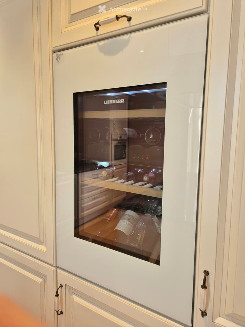 A modern white kitchen cabinet with a built-in wine storage unit featuring glass doors and multiple shelves holding various wine bottles.
