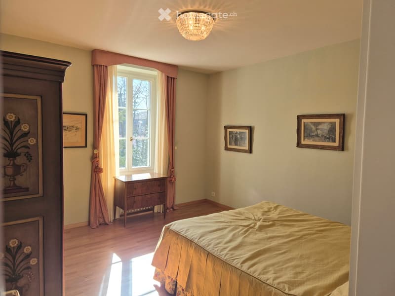 a bedroom with a double bed, wooden dresser, window with curtains, wooden floor, and framed pictures on the wall