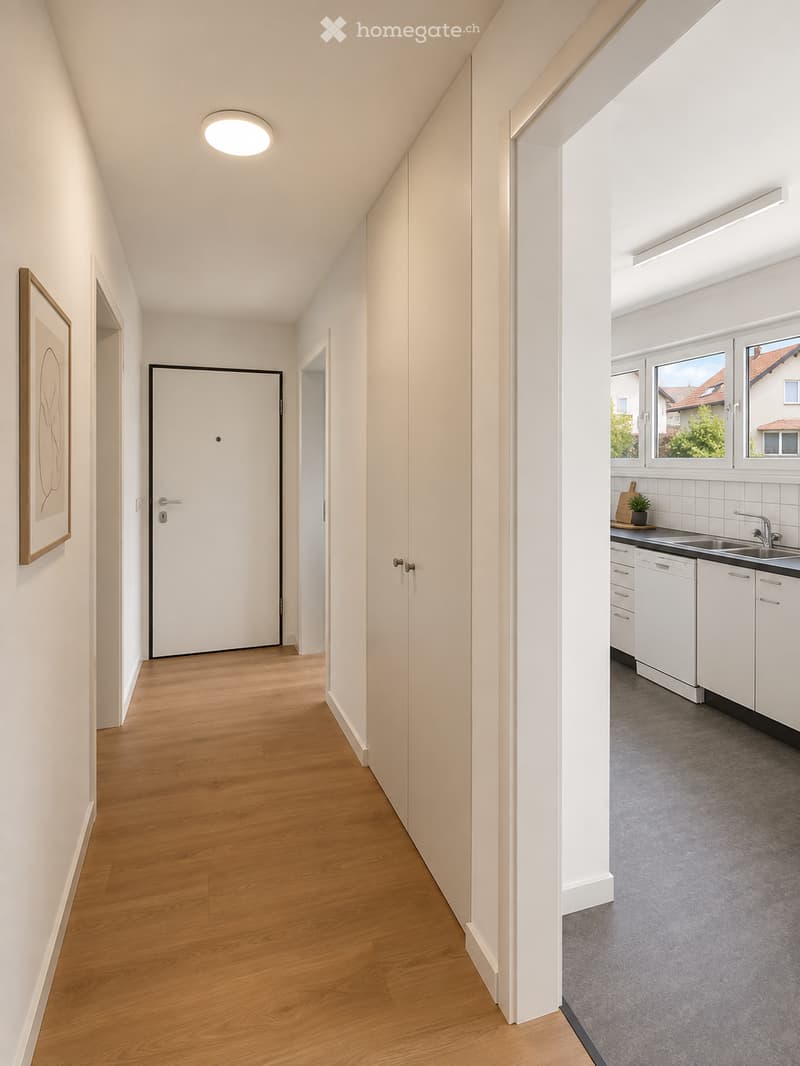 Bright hallway with wooden flooring, white walls, and a recessed ceiling light. A door leads to a kitchen area with white cabinets, a sink, and a dishwasher.