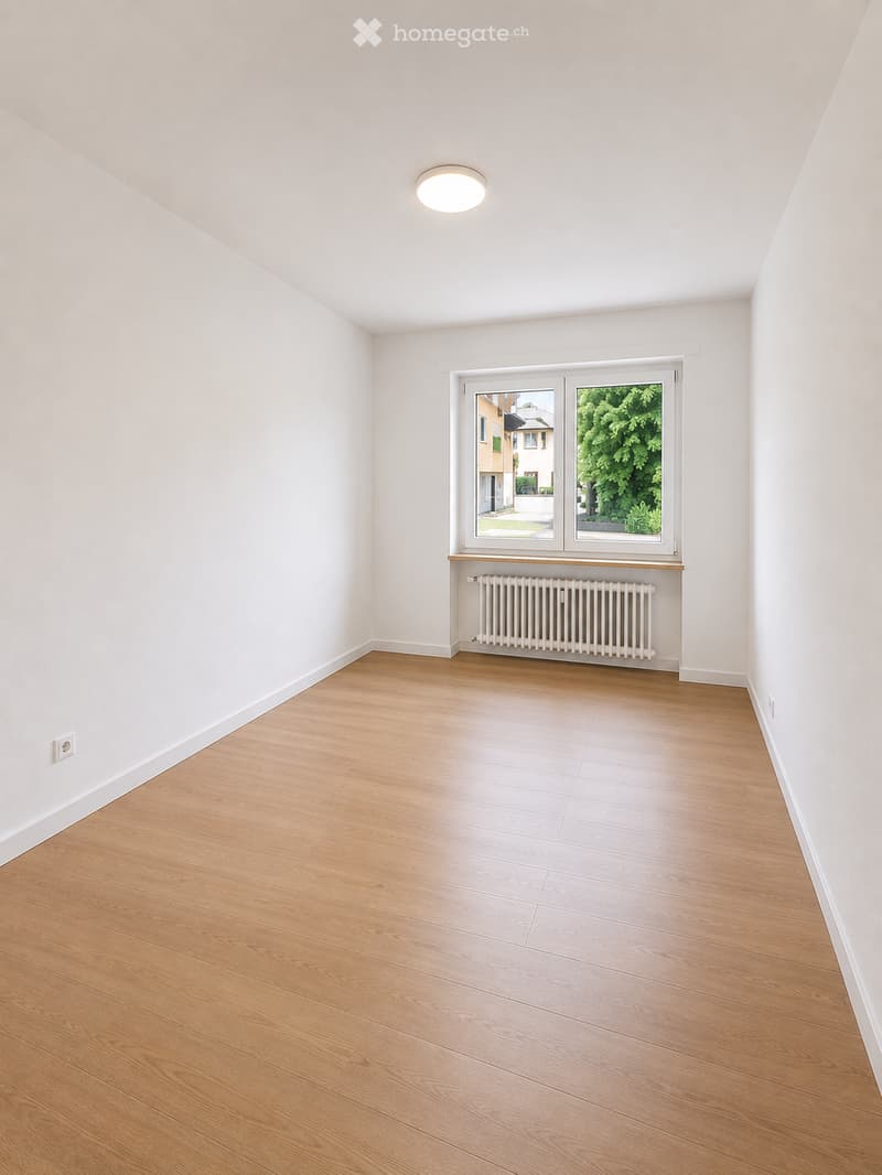 Empty room with light wooden flooring, white walls, a large window with a view of greenery, a radiator, and a ceiling light fixture.