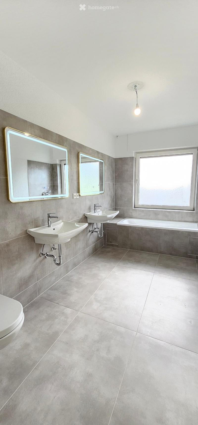 Modern bathroom with two sinks, mirrors, a bathtub, and a toilet, featuring grey tiles and a large window.
