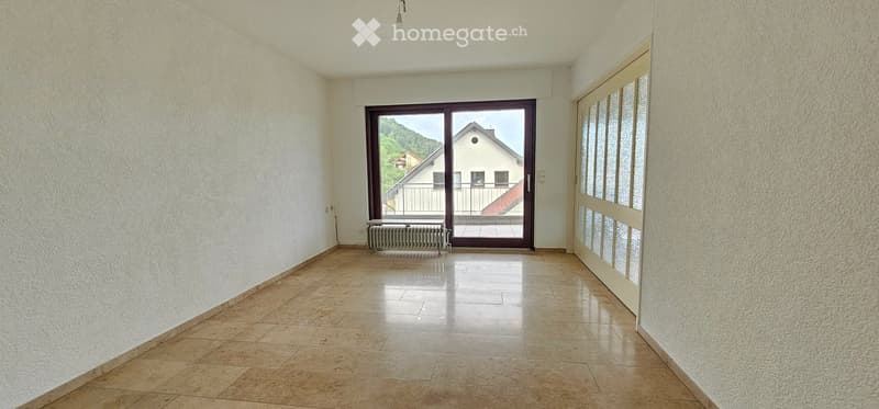 Empty room with tiled floor, white walls, large sliding glass door leading to a balcony, radiator, and a view of a house and greenery outside.